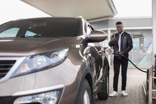 Happy berjanggut pria Afrika memberi jempol dan tersenyum sambil mengisi bahan bakar mobil mewah modern di pompa bensin. Seorang pria di pompa bensin mengisi mobilnya dengan bensin — Stok Foto