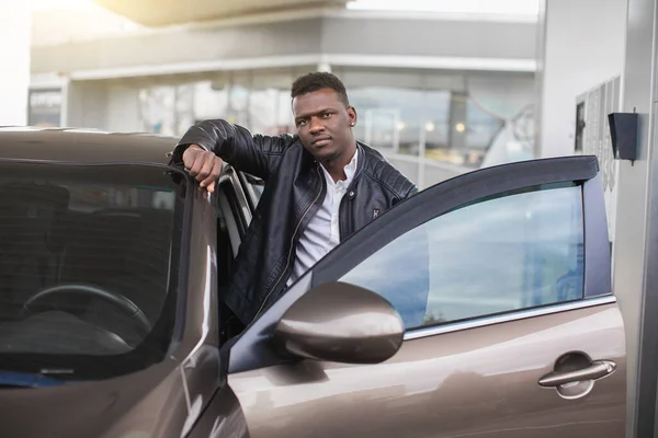 Station essence, concept de ravitaillement en voiture. Homme africain positif en tenue décontractée appuyé sur sa porte de voiture et regardant la caméra tout en se tenant debout sur fond flou de la station-service — Photo