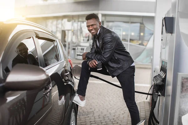 Feliz joven afroamericano guapo en una gasolinera, apoyándose en su coche de lujo y bebiendo café para llevar mientras repostan un coche —  Fotos de Stock