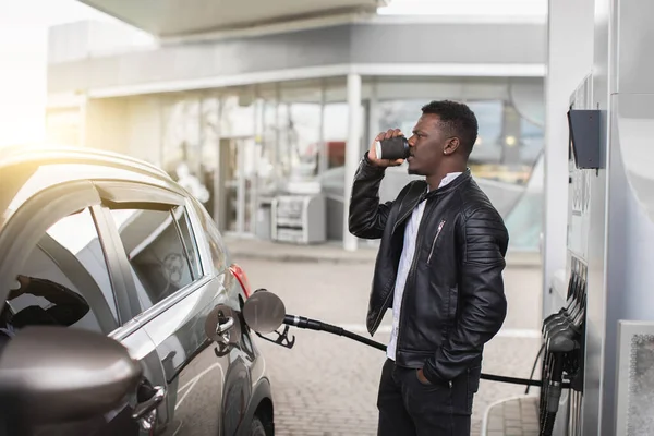 Giovane bel ragazzo africano, uomo d'affari in abito casual nero, facendo rifornimento della sua auto moderna al distributore di benzina e bevendo caffè per andare — Foto Stock