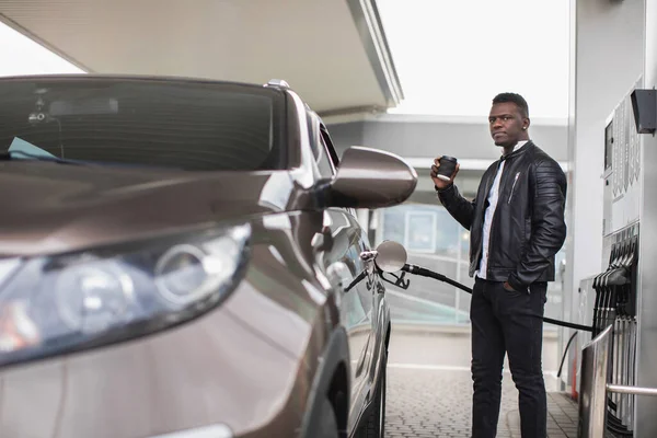 Νεαρός Αφροαμερικανός επιχειρηματίας σε casual μαύρο ντύσιμο, κοιτάζοντας μακριά, ενώ πίνοντας καφέ για να πάει, ανεφοδιάζοντας το σύγχρονο πολυτελές αυτοκίνητό του στο βενζινάδικο. — Φωτογραφία Αρχείου