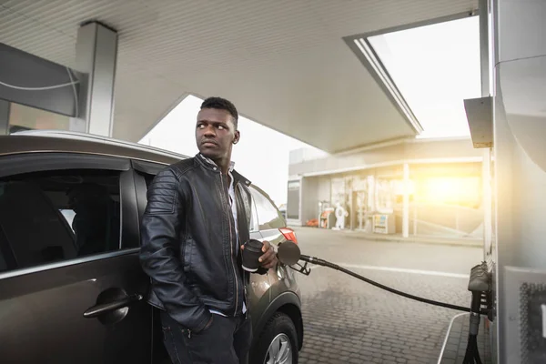 Έννοια βενζινάδικου. Χαλαρός ονειρεμένος νεαρός όμορφος Αφρικανός επιχειρηματίας με μαύρα ρούχα, απολαμβάνοντας τον καφέ του, ενώ ανεφοδιάζει το πολυτελές αυτοκίνητό του στο βενζινάδικο — Φωτογραφία Αρχείου