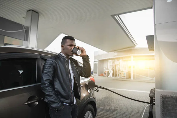 Ung fokuserad afrikansk affärsman i svart läder jacka, dricka sitt kaffe för att gå, medan du står lutad på bilen vid den moderna bensinstationen — Stockfoto