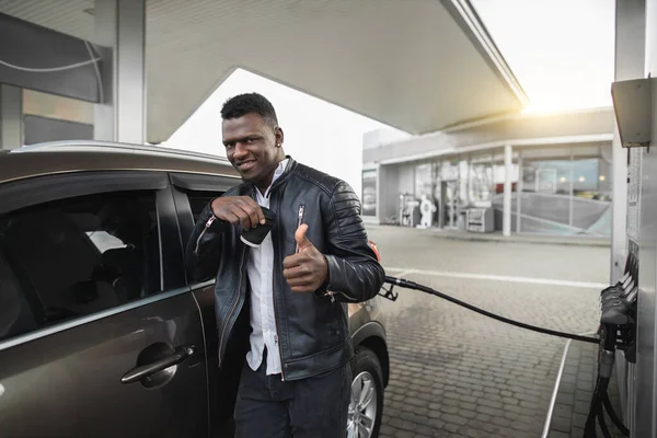 Πορτρέτο του νεαρού όμορφος χαμογελαστός Αφροαμερικανός, φορώντας μαύρα casual ρούχα, κοιτάζοντας κάμερα με καφέ για να πάει, δείχνοντας τον αντίχειρα προς τα πάνω, ενώ ανεφοδιασμό αυτοκινήτων στο βενζινάδικο — Φωτογραφία Αρχείου