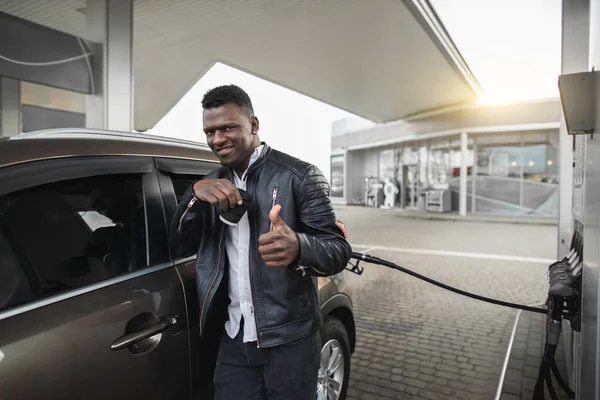 Retrato del joven guapo y sonriente hombre afroamericano, vestido con ropa casual negra, mirando a la cámara con café para llevar, mostrando el pulgar hacia arriba, mientras repostaba automóvil en la gasolinera —  Fotos de Stock