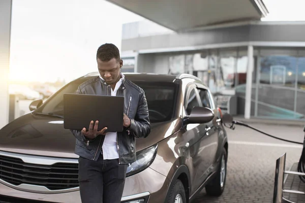 Pengusaha muda Afrika-Amerika, berdiri di depan mobilnya dan bekerja pada laptop pc, sambil mengisi bahan bakar mobil di pompa bensin. Blurred pom bensin di latar belakang — Stok Foto