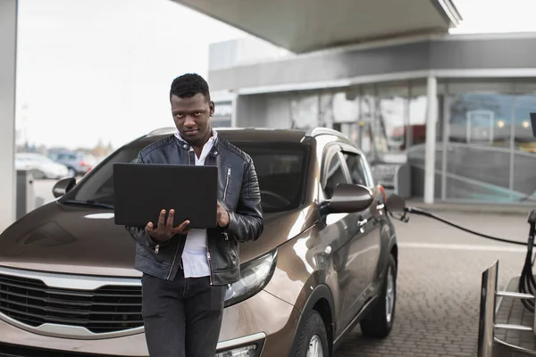 Jeune homme d'affaires afro-américain confiant, debout devant sa voiture et travaillant sur ordinateur portable, tout en ravitaillant l'automobile à la station-service. Station-service floue en arrière-plan — Photo