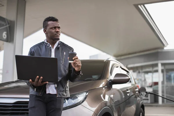 Μαύρος επιχειρηματίας στο πρατήριο βενζίνης, στέκεται κοντά στο αυτοκίνητο με take away καφέ και φορητό υπολογιστή και κοιτάζοντας μακριά, ενώ ανεφοδιάζει το αυτοκίνητό του. Βενζινάδικο και άνθρωποι έννοια — Φωτογραφία Αρχείου