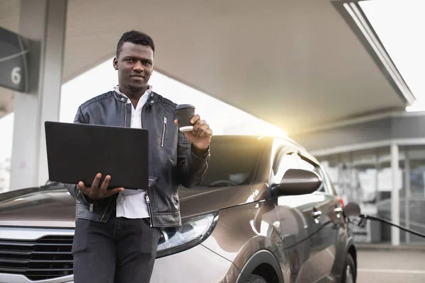 Rifornimento auto, affari, concetto di persone. Ritratto di bel giovane africano in abito casual nero, in posa per la fotocamera con computer portatile e caffè da andare, mentre la sua auto sta facendo rifornimento alla stazione di servizio — Foto Stock