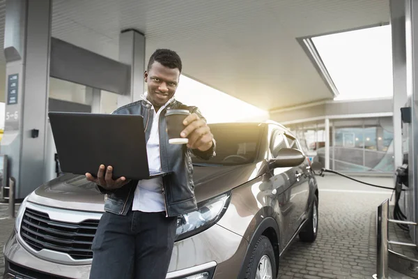 Pengisian bahan bakar mobil, bisnis, konsep orang. Potret tampan pemuda Afrika berpakaian hitam santai, berpose ke kamera dengan laptop dan kopi untuk pergi, sementara mobilnya mengisi bahan bakar di pompa bensin — Stok Foto