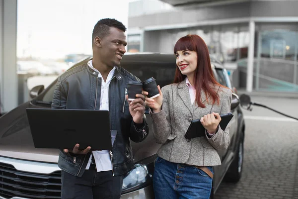 Werk en pauze op de weg. Portret van lachende jonge multiraciale man en vrouw, zakenpartners of koppel, vullen moderne auto op benzinestation, klinkende papieren glazen met take away koffie — Stockfoto