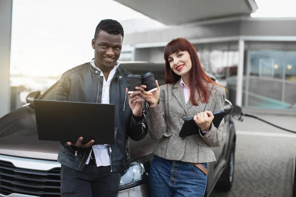 Selektivt fokus av glada multietniska par, poserar till kameran med leende, klinkande pappersglas med kaffe att gå, när du står vid bensinstationen, tankning den moderna bilen — Stockfoto