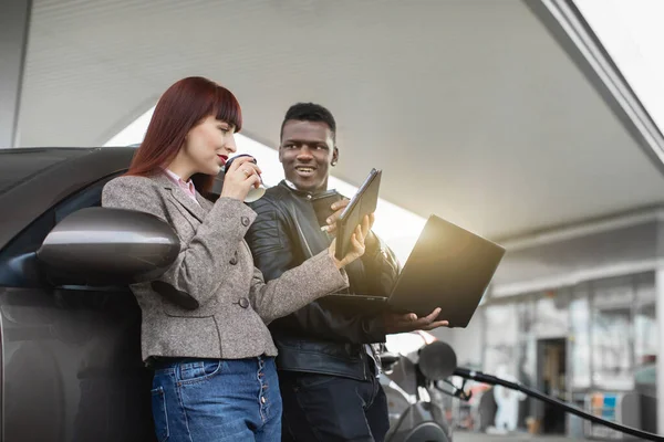 Heureux couple multiethnique ou collègues d'affaires, homme africain et femme blanche tenant des tasses en papier avec café et en utilisant tablette et ordinateur portable pc, tout en ravitaillement voiture à la station-service — Photo