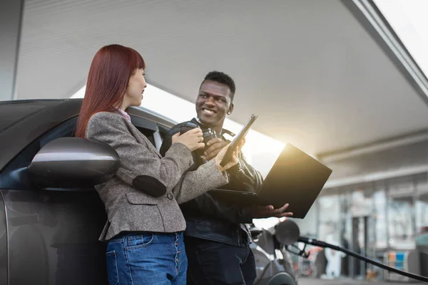 Gros plan horizontal du couple multiracial riant, appuyé sur une voiture de luxe, parlant entre eux, tenant tablette et ordinateur portable, buvant du café à emporter, ravitaillant la voiture à la station-service — Photo