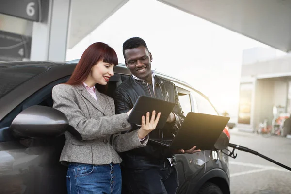 Bekerja dan istirahat di pom bensin. Shot of young African man and pretty Caucasian lady, showing each other smth on digital tablet, while standing together, fuel the car at gas station — Stok Foto