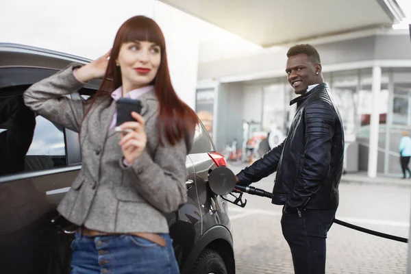 Handsome smiling African man, fueling luxury car at petrol station, while woman with take away coffee stands in front of him, leaning on the auto. Focus on black man