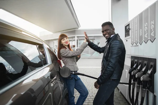 Ett par på macken. Glada glada multiracial par vänner eller kollegor, poserar inför kameran, har kul samtidigt tanka lyxbil på bensinstationen, ger high five varandra och ler — Stockfoto