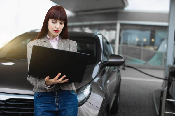 Benzinestation, bedrijf en mensen concept. Jonge mooie zakenvrouw op tankstation, terwijl het vullen van haar auto, geconcentreerd en werken op laptop, leunend op de auto — Stockfoto