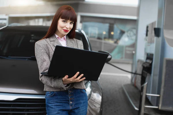 Närbild porträtt av vackra kaukasiska affärskvinna, lutar sig på sin bil medan tankning den på bensinstationen, arbetar på laptop dator dator och ler mot kameran — Stockfoto
