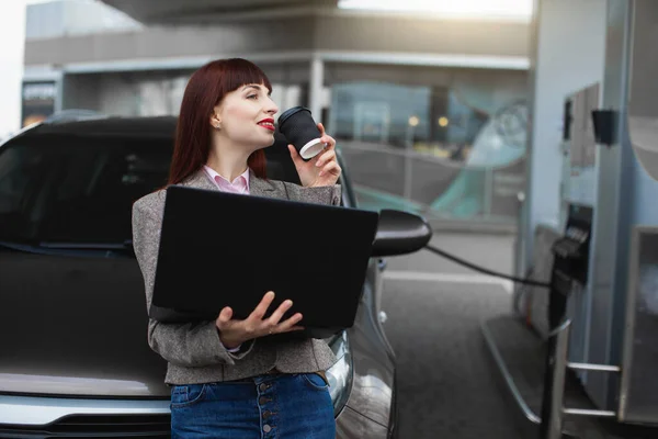 Pengusaha wanita di pom bensin. Wanita muda cantik menggunakan laptop pc dan minum kopi untuk pergi sambil mengisi bahan bakar mobilnya di pompa bensin — Stok Foto