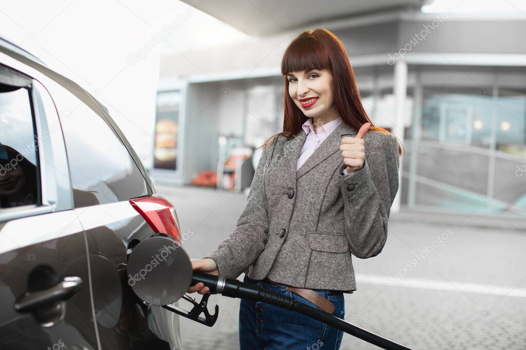 Attractive cheerful young business woman, smiling and showing her thumb up to camera, while refueling her luxury car at gas station