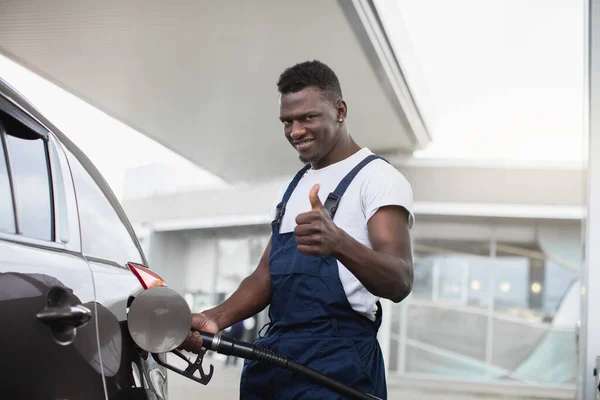 Gülümseyen Afrikalı Amerikalı adam, benzin istasyonunda çalışan, beyaz tişört ve mavi tulum giyen, baş parmağını gösteren, lüks bir arabayı dolduran — Stok fotoğraf
