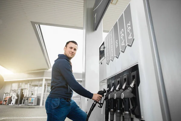 Joven hombre guapo en ropa casual, repostando su coche con gasolina en una gasolinera, tomando el arma de llenado del panel de gasolina — Foto de Stock