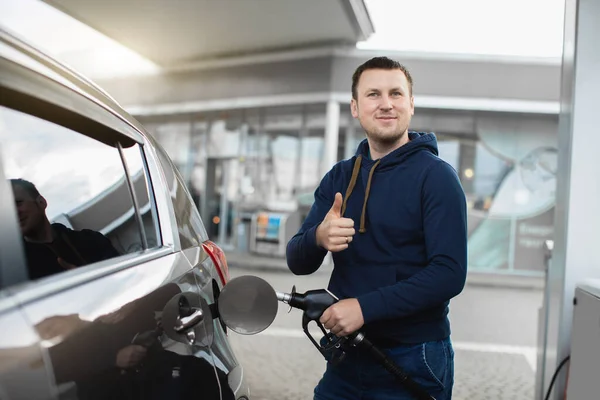 Joven hombre caucásico sonriente repostando su coche moderno en una gasolinera, posando ante la cámara con el pulgar hacia arriba —  Fotos de Stock