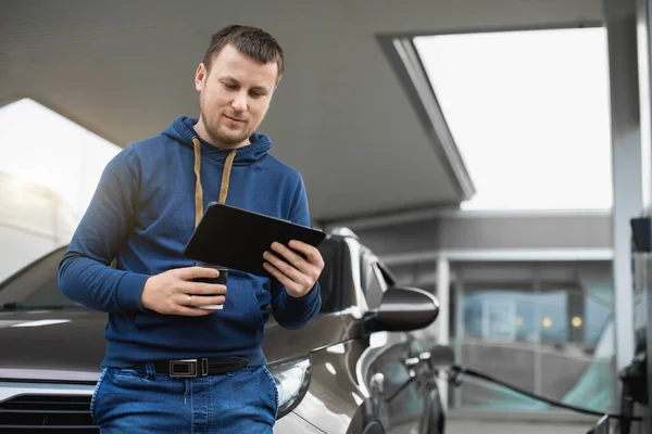 Pemuda tampan Kaukasia, berdiri di depan mobilnya dan bekerja di tablet pc, sambil mengisi bahan bakar mobil di pompa bensin. Blurred pom bensin di latar belakang — Stok Foto