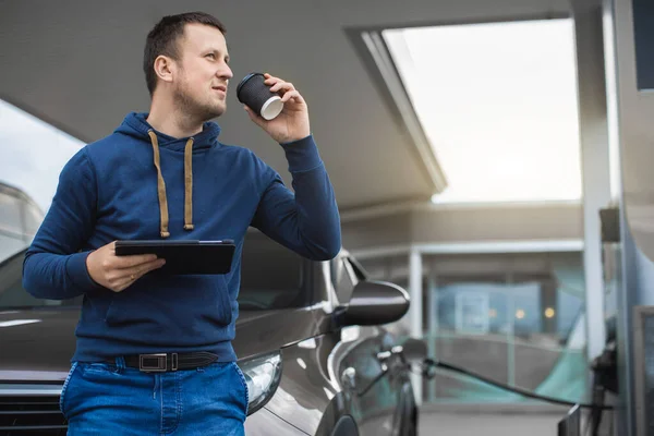 Όμορφος νεαρός καυκάσιος άνδρας στο βενζινάδικο, στέκεται κοντά στο αυτοκίνητο με take away καφέ και tablet pc και κοιτάζοντας μακριά, ενώ ανεφοδιάζει το αυτοκίνητό του. Βενζινάδικο και άνθρωποι έννοια — Φωτογραφία Αρχείου