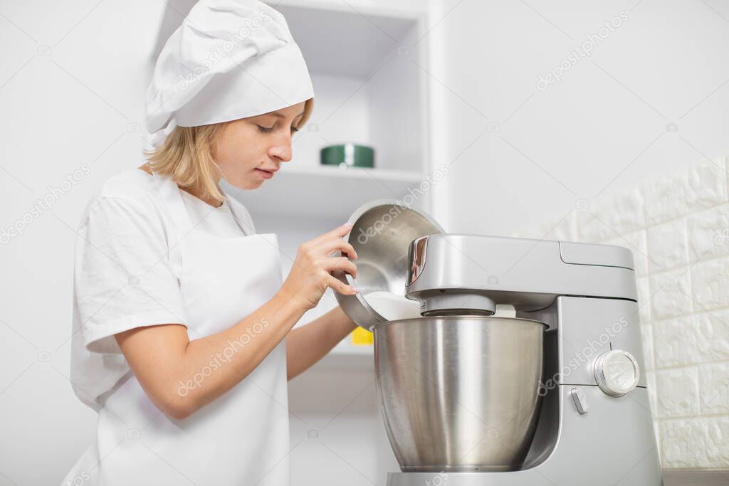 Young blond girl, professional confectioner, wearing white apron and chefs hat, cooking in cozy light kitchen interior, using mixer or food processor for whipping egg whites