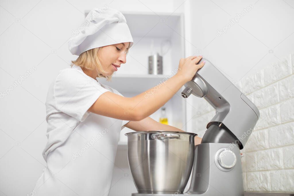 Close up indoor shot of young blond girl confectioner, wearing in white apron and chefs hat, using stand modern food processor for mixing dough or baking ingredients