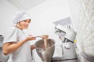 Kenar manzaralı genç kız şekerleme şefinin fotoğrafı. Karışık hamuru kontrol ediyor, elinde bir spatula tutuyor, hamur işini metal bir kasede yapıyor.