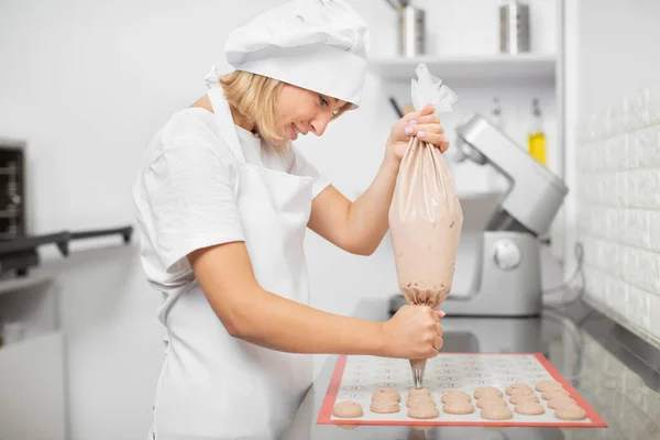 Sorridente giovane chef donna, godendo il suo lavoro in pasticceria, fare i maccheroni, spremere l'impasto da una siringa di cottura su una teglia da forno. Preparazione di dessert francese — Foto Stock