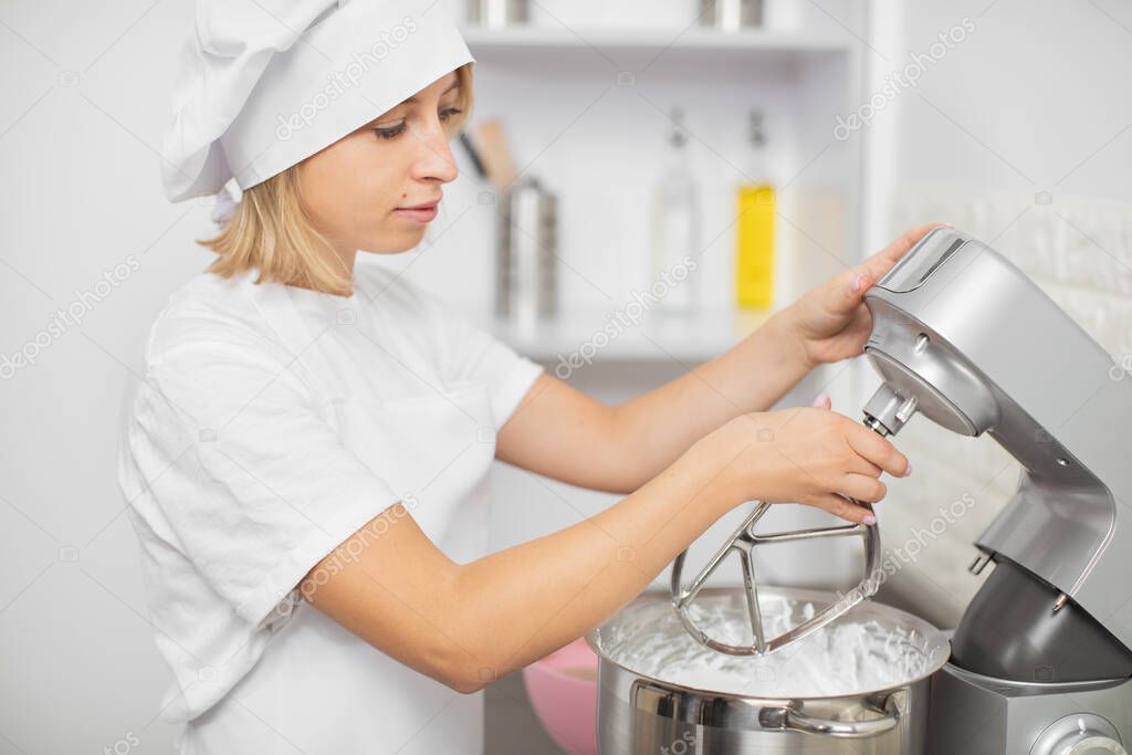 Pretty young blond girl confectioner, using modern electric food processor to mixing the dough for baking a cake or cookies in light kitchen interior