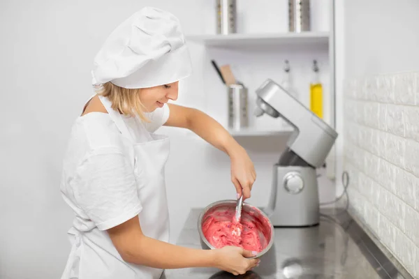 Hübsche junge Frau, professionelle Konditorin, bereitet rote Sahne oder Teig in einer Edelstahlschüssel zu und mischt sie mit dem Spachtel. In der Küche backen, Sahne für Kuchen oder Macarons zubereiten — Stockfoto