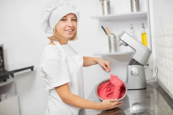 Giovane bella donna bionda, indossa un cappello bianco chef e grembiule bianco, mescolando deliziosa crema di salsa rossa in ciotola con spatola, in posa per la fotocamera con sorriso in interni cucina luce — Foto Stock