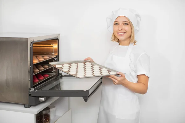 Confectionery shop, macarons baking. Pretty woman confectioner in white uniform and hat, holds a batch of macarons on baking tray, ready to put it into the oven.