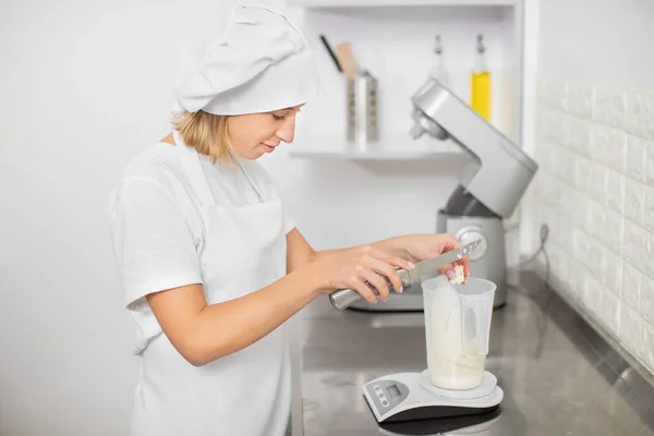 Formaggio in cottura o al forno. Ritratto ravvicinato di graziosa giovane cuoca dolciaria, in cappello e grembiule, tagliato con un coltello gustoso formaggio gorgonzola nel misurino sui pesi della cucina — Foto Stock