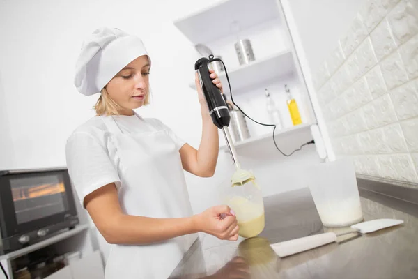 Giovane donna, panettiere professionista, mescola ingredienti per ganache crema nel misurino, tenendo in mano un frullatore elettrico sommergibile. Lavorare in pasticceria, fare dolci — Foto Stock