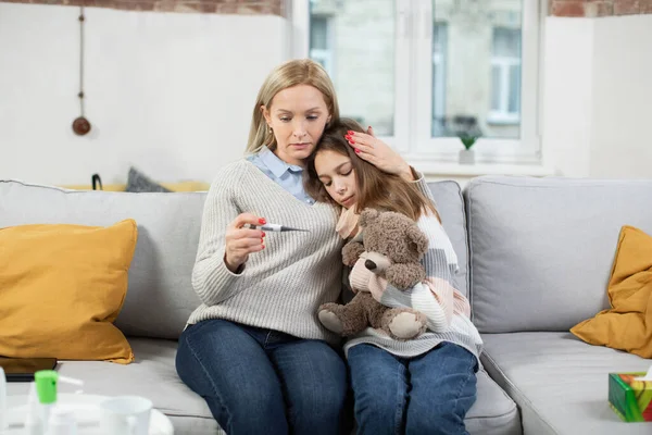 Orta yaşlı endişeli sarışın kadın, anne, evdeki kanepede oturuyor, hasta ergen kızına sarılıyor, ölçtükten sonra termometreden ısıyı kontrol ediyor. Grip, soğuk algınlığı, virüs enfeksiyonu. — Stok fotoğraf