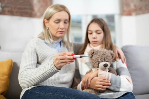 Mature blond woman, checking her sick daughters temperature on thermometer after measuring. Sad ill girl, holding teddy bear, looks at thermometer with her mom. Focus on thermometer — Stock Photo, Image