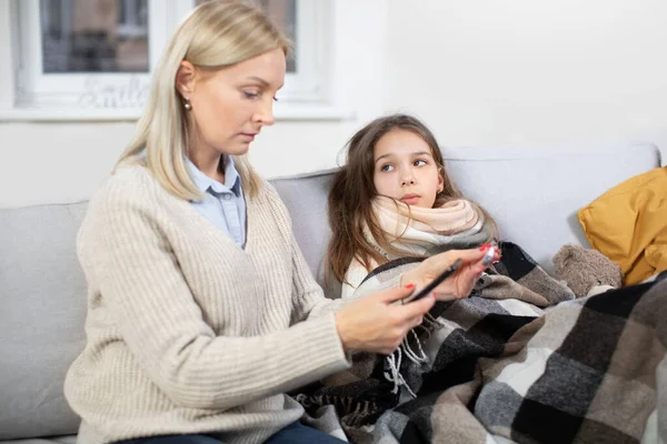 Ziek kind. Schattig tienermeisje, liggend op de bank thuis met sjaal om de nek, bedekt met deken en kijkend naar haar moeder, de temperatuur controleren en de dokter bellen. Focus op ziek meisje — Stockfoto