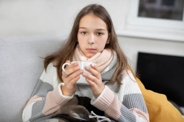 Home self-treatment. Medical quarantine. Flu, cold, covid-19 therapy. Sad tired ill teen girl wrapped in a blanket, sitting on sofa at home, holding cup of hot tea or medicine and looking at camera