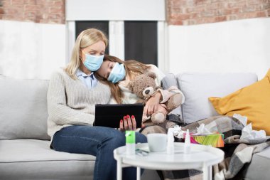 İzolasyon, hastalık, enfeksiyon, koronavirüs ve karantina konsepti. Koruyucu maskeli hasta anne ve kız çocuğu oturma odasındaki kanepede birlikte oturup tablet bilgisayar kullanıyorlar.