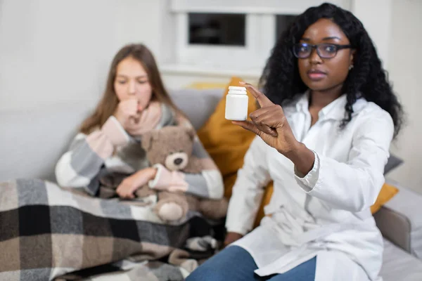 Patient and doctor. Female African doctor, visiting her sick teen girl patient at home for check up, showing to camera white box with pills for colf and flu treatment. Focus on box with tabs