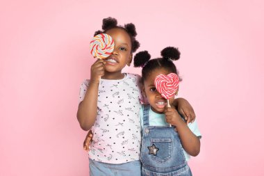 İki tatlı Afrikalı kızın gözlerini renkli lolipoplarla kaplarken, pembe arkaplanda kameraya gülümserken çekilen yatay çekimini kapat.