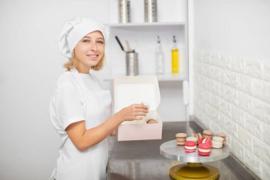 Genç, gülümseyen çekici kadın şekerlemecinin yatay görüntüsü. Pasta dükkanının mutfağında dururken taze rengarenk makaronları şimdiki kutuya koyuyor.