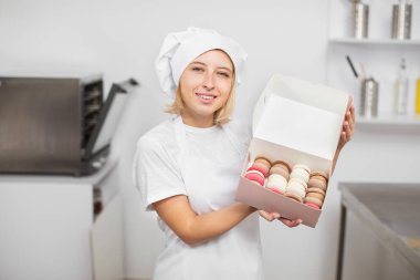 Şekerleme atölyesinde çalışıyorum. El yapımı badem kurabiyeleri. Gülümseyen genç kadın pasta şefi, kameraya lezzetli taze makaronlarla hediye kutusunu gösteriyor.