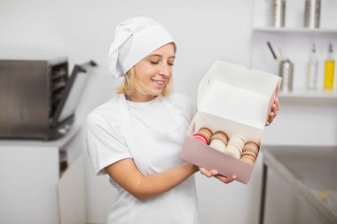 Beyaz şapkalı, önlüklü, gülümseyen genç kız pastacı. Mutfağın içinde duruyor. Lezzetli kurabiyelerle dolu bir kutu tutuyor.